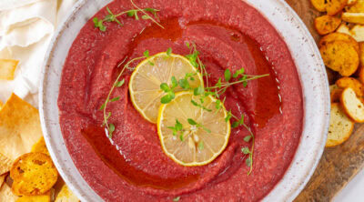 square crop of pink colored beet hummus with slices of lemon garnish in a white bowl with crackers and pita chips around.