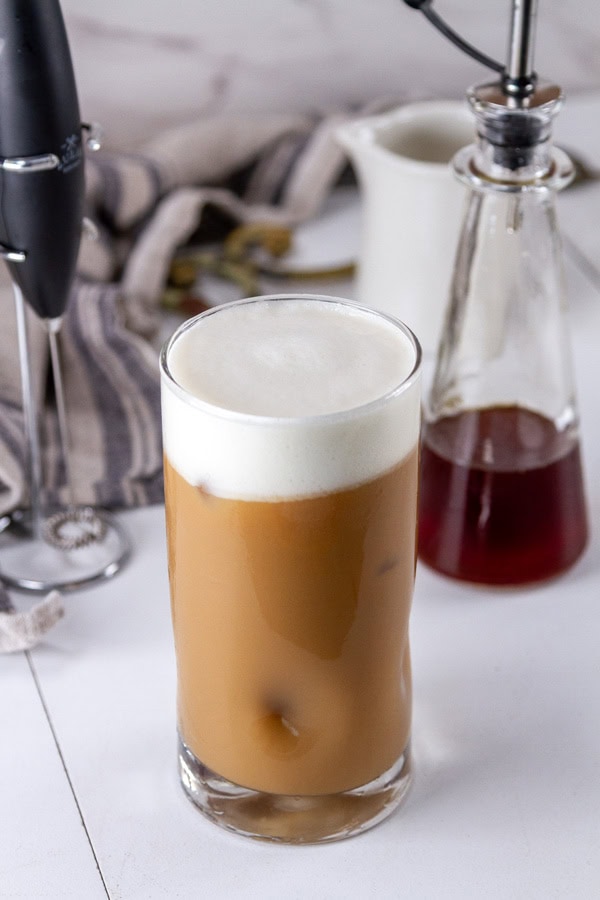 a chai latte with cold foam in front of a frother and the jar of syrup.