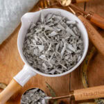 square crop of a white measuring cup filled with dried crumbled sage on a wooden background.