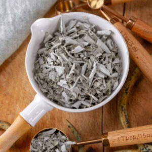 square crop of a white measuring cup filled with dried crumbled sage on a wooden background.
