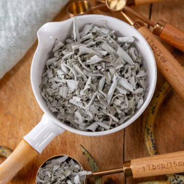 square crop of a white measuring cup filled with dried crumbled sage on a wooden background.