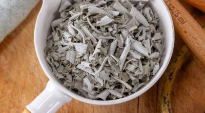 square crop of a white measuring cup filled with dried crumbled sage on a wooden background.