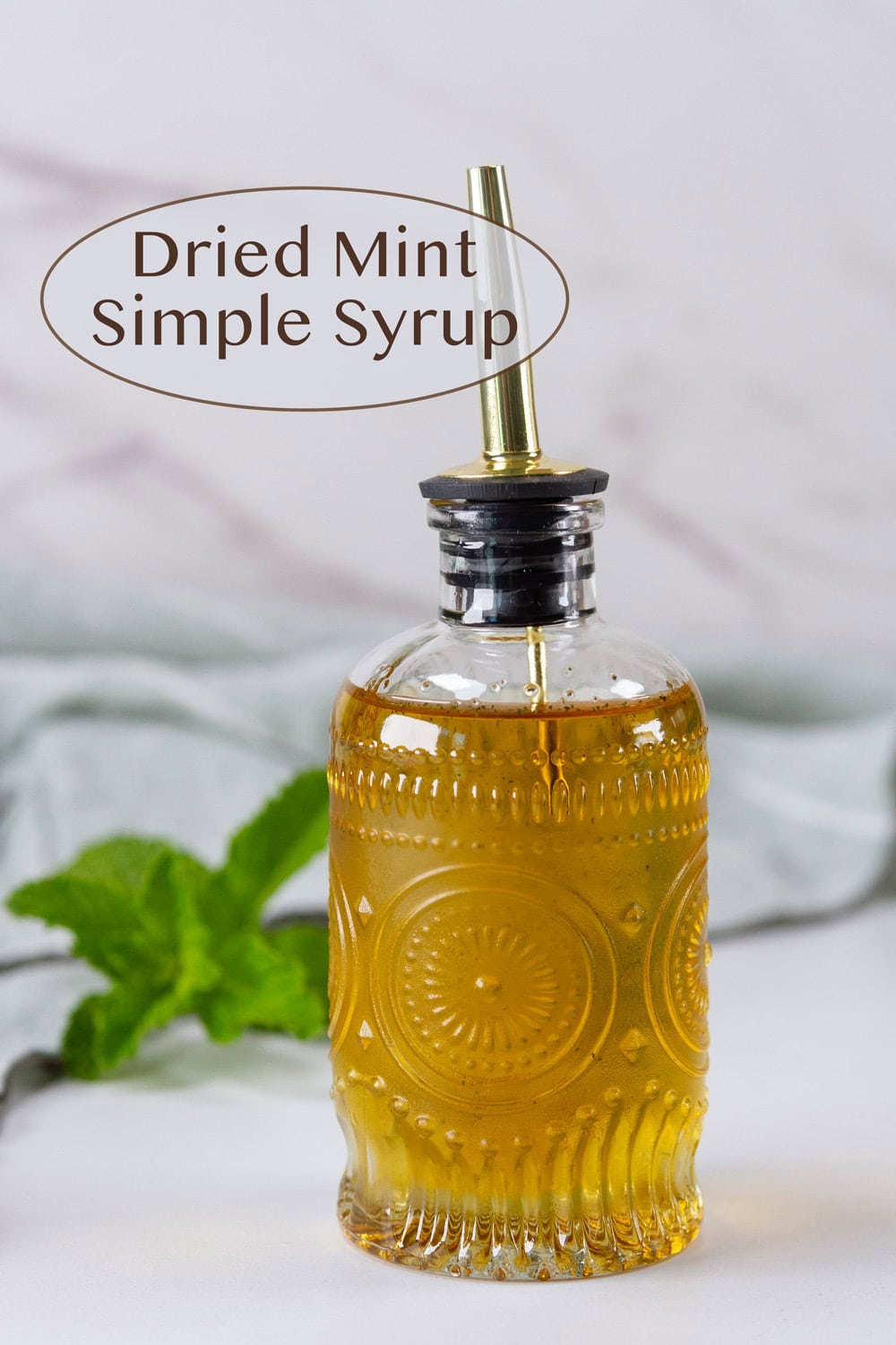a glass dispenser bottle filled with mint simple syrup and a mint sprig behind it.