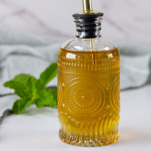 square crop of a glass dispenser bottle of simple syrup with a mint sprig behind it.