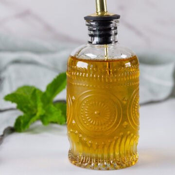 square crop of a glass dispenser bottle of simple syrup with a mint sprig behind it.