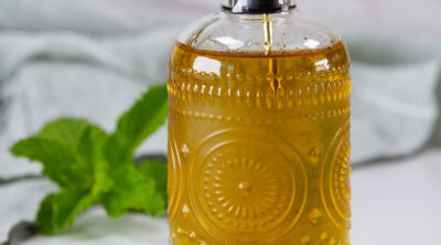 square crop of a glass dispenser bottle of simple syrup with a mint sprig behind it.