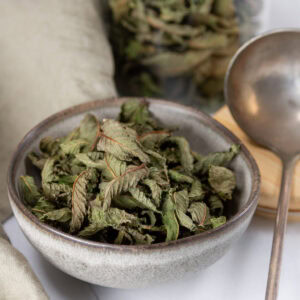 square crop of a grey bowl filled with dried mint and a silver spoon to the right.