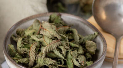 square crop of a grey bowl filled with dried mint and a silver spoon to the right.