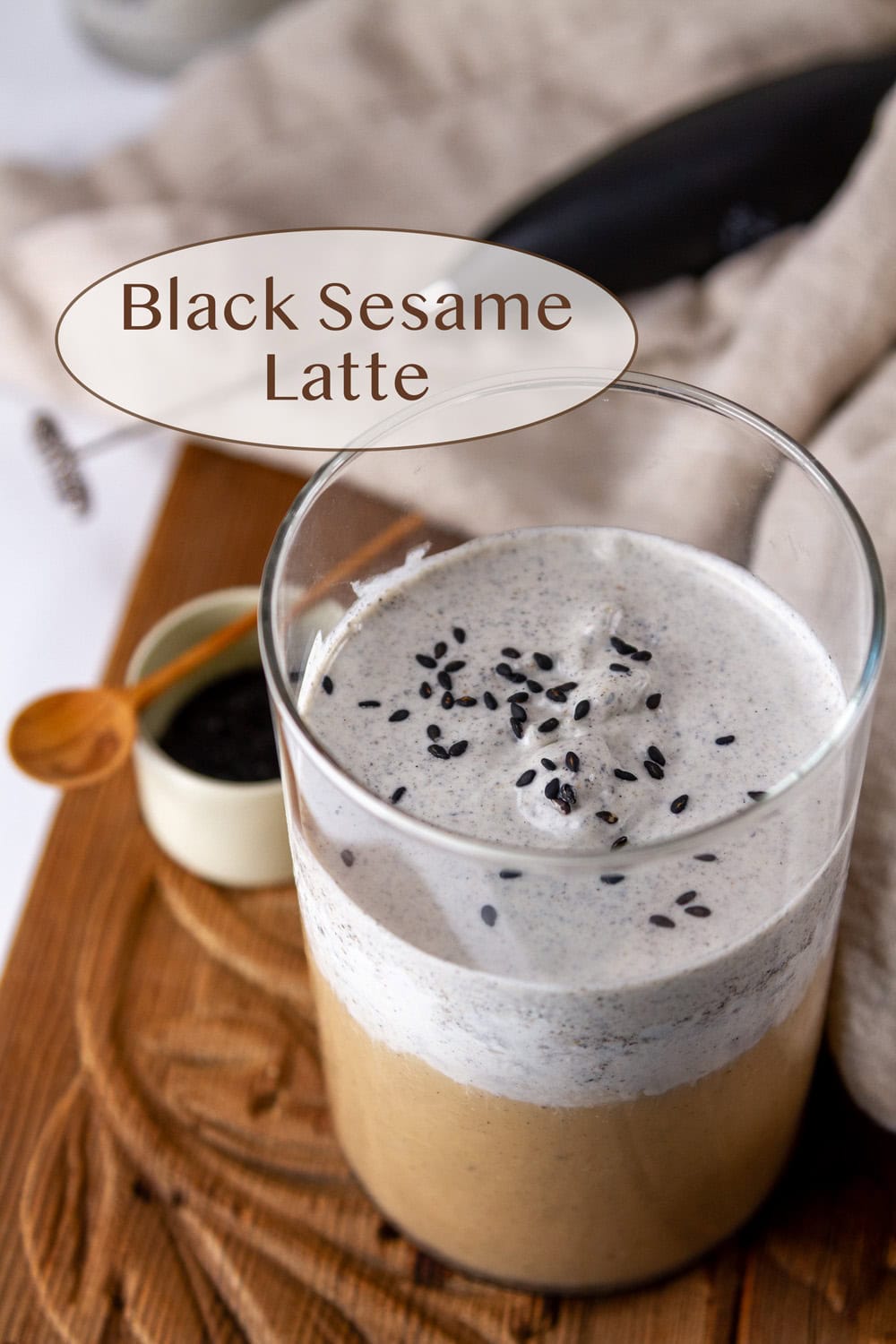 an Iced Black Sesame Latte with black sesame cold foam on a wooden serving board with a cloth and a small dish of black sesame powder.
