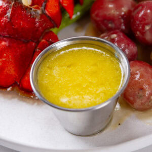 square crop of a small ramekin of butter sauce next to potatoes and lobster.