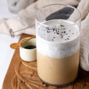 square crop of a latte with cold foam on a wooden tray with a dish of black sesame powder.