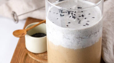 square crop of a latte with cold foam on a wooden tray with a dish of black sesame powder.