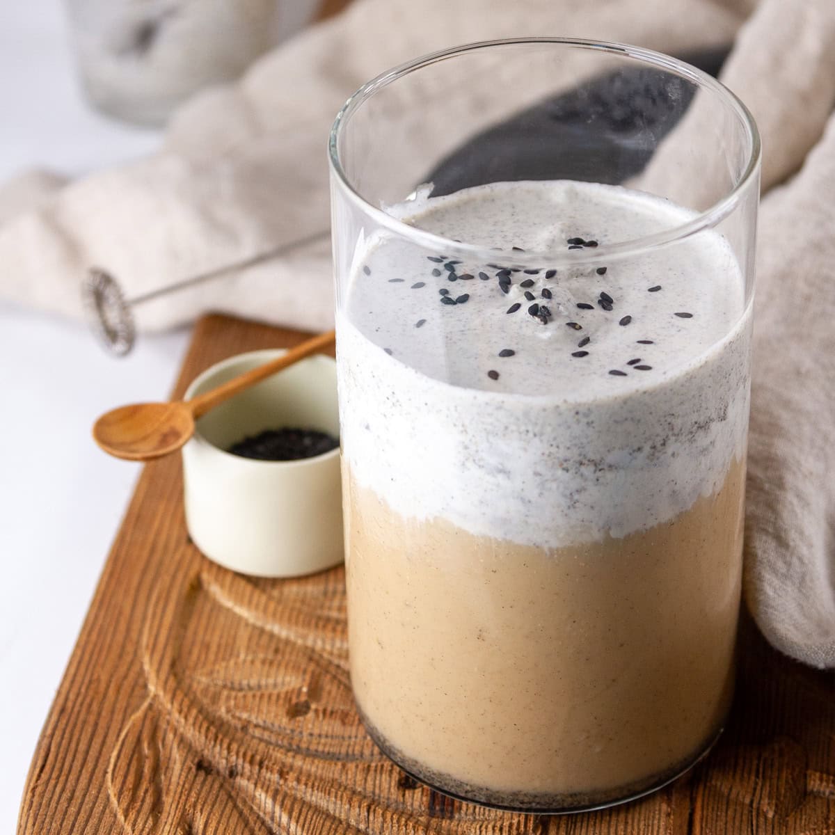 square crop of a latte with cold foam on a wooden tray with a dish of black sesame powder.