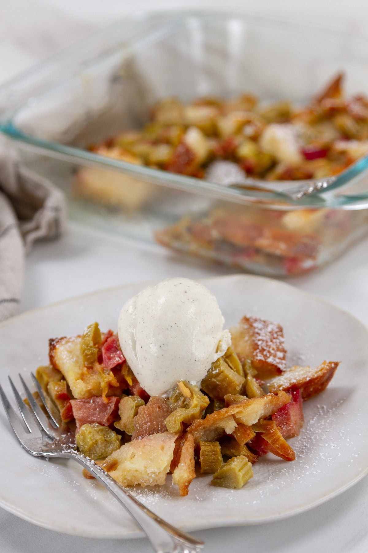 a serving of rhubarb bread pudding with vanilla ice cream in front of the baking dish full of the dessert.