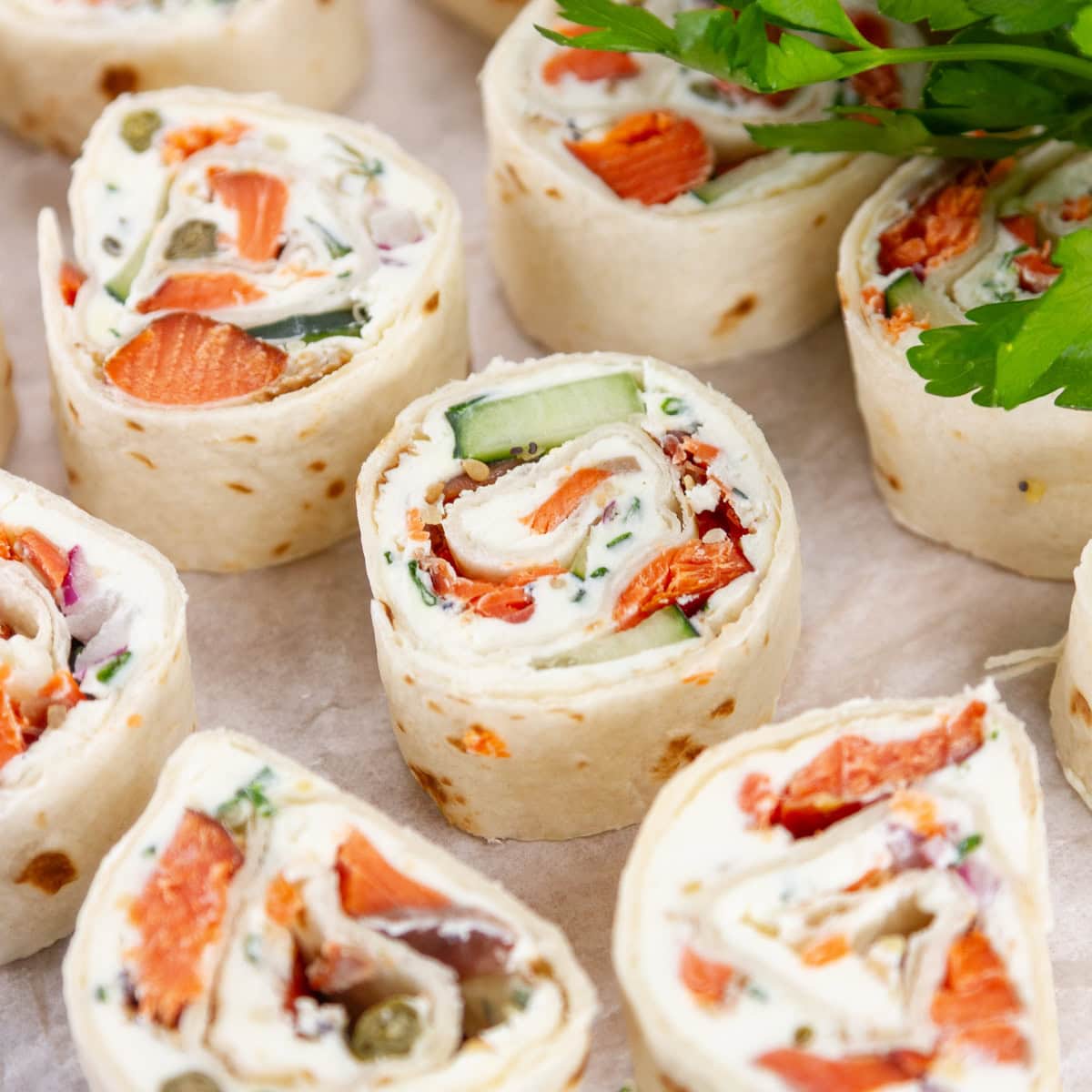 square crop of several pinwheel appetizers filled with cream cheese and salmon, and there's fresh parsley in the upper right.