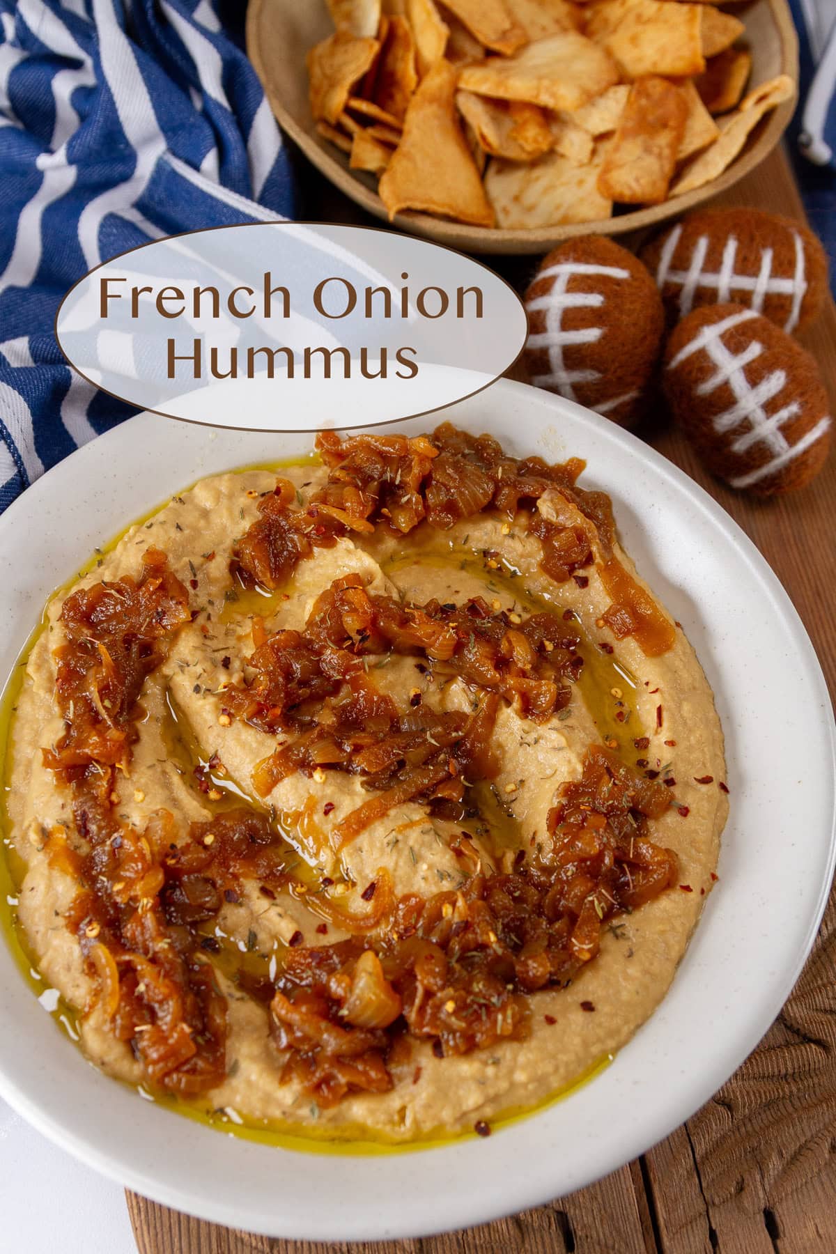 a white bowl filled with hummus topped with onions, some footballs to the upper right and a bowl of pita chips.