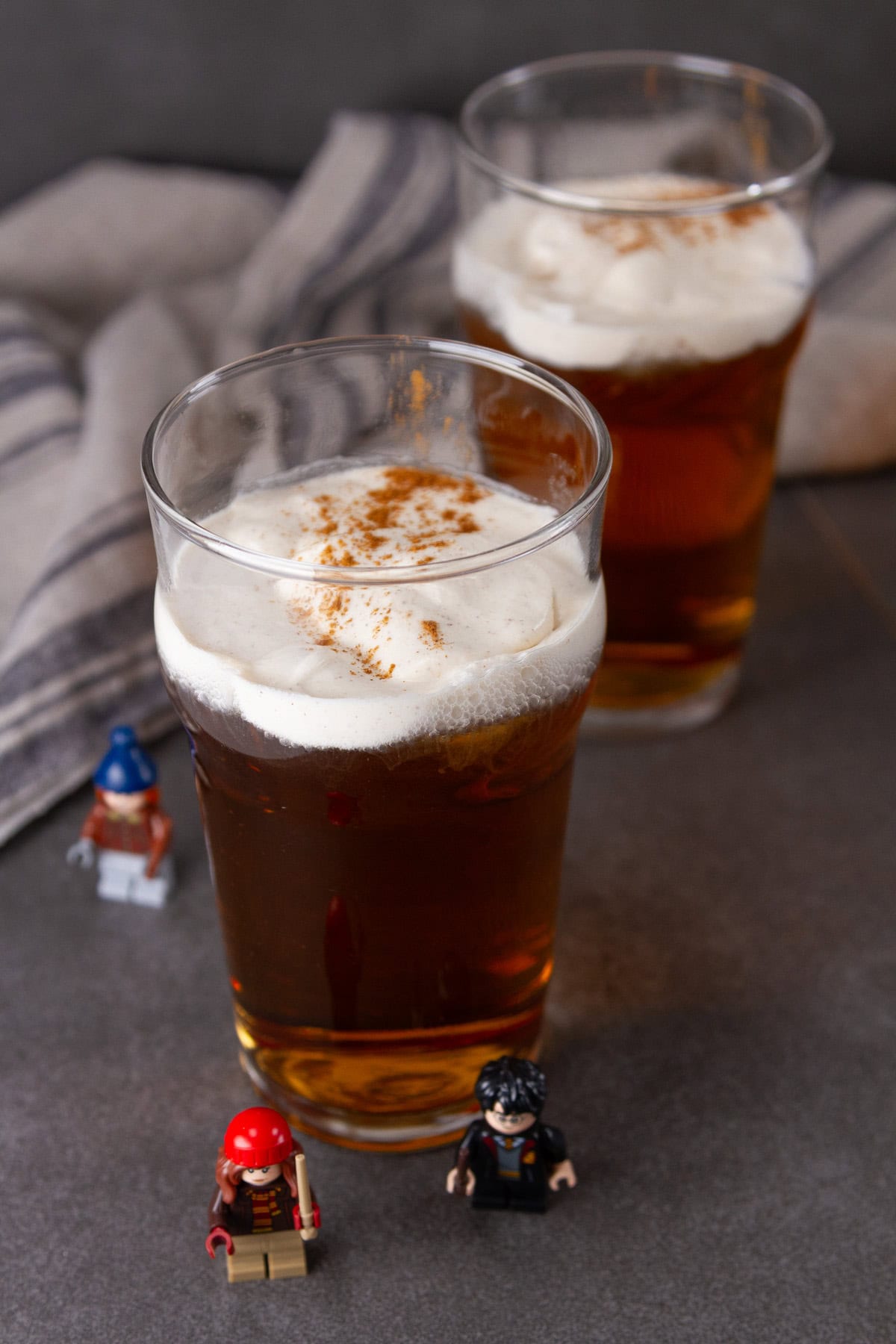 two butterbeers on a dark background with striped cloth in the back and Harry Potter figures at the bottom.
