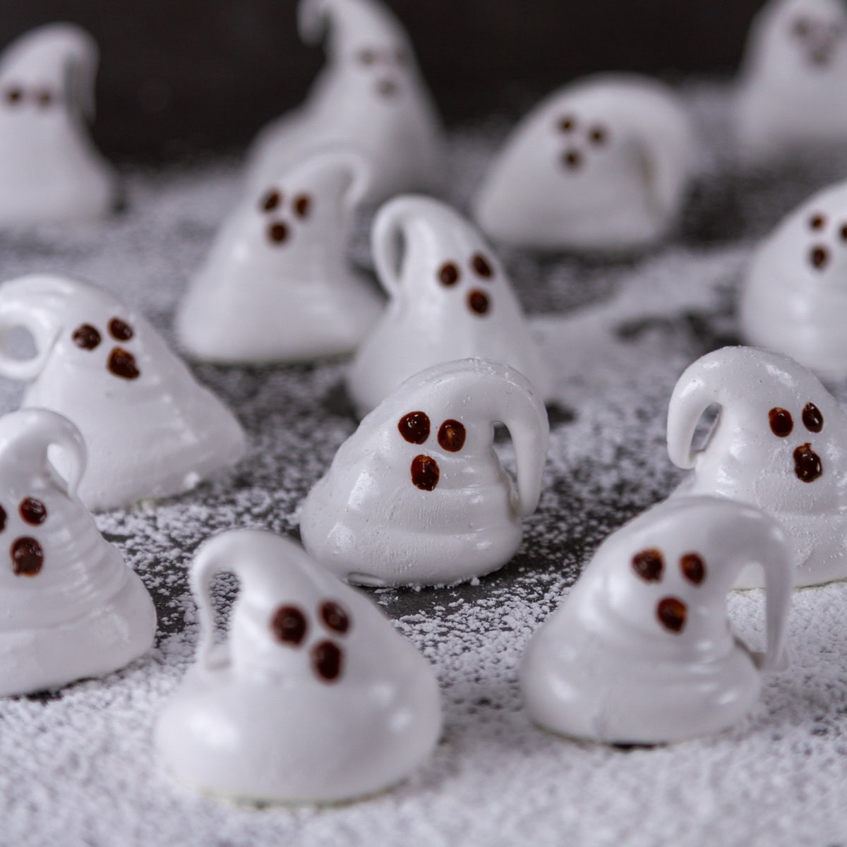 square crop of a bunch of ghost shaped marshmallows on powdered sugar on a black background.